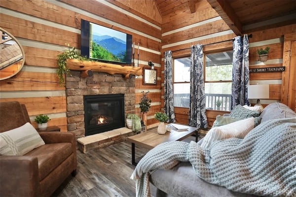 Log cabin living room with stone fireplace, leather seating, beds with knit blankets, and mountain view through windows