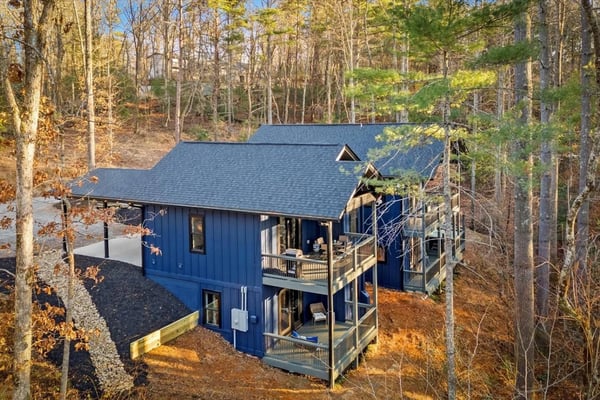 Modern blue cabin with gray roof nestled among bare and evergreen trees on hillside, featuring wraparound deck and driveway.