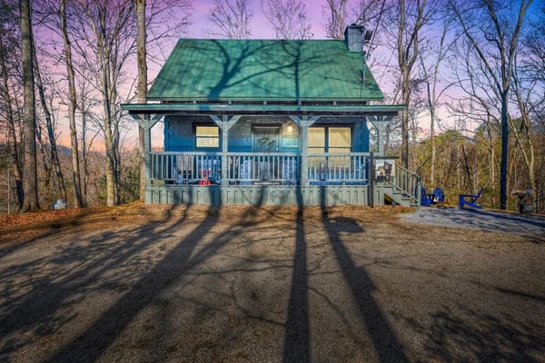 Blue cottage with green metal roof surrounded by bare trees at dusk, front porch with white railings and Adirondack chairs visible.
