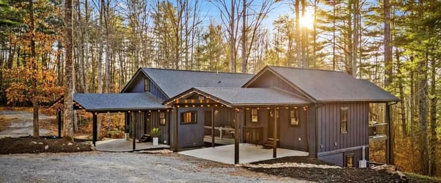Modern dark wood cabin with wraparound porches nestled among tall pine trees at sunset