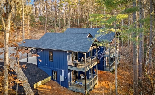 Modern blue cabin with gray roof nestled among bare and evergreen trees on hillside, featuring wraparound deck and gravel driveway.